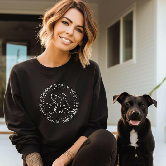 Woman wearing a crewneck sweatshirt sitting beside a small black dog. Her comfy and oversized black sweatshirt has a graphic of a dog and a cat with a positive message.