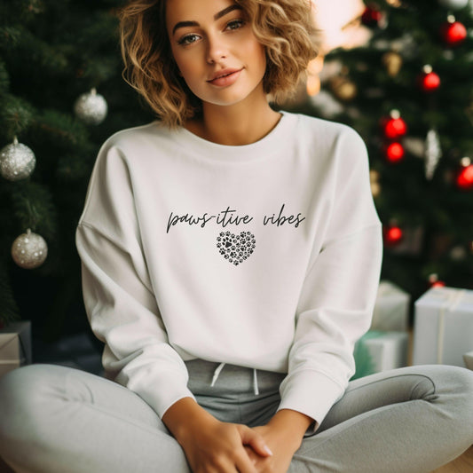 Woman sitting cross legged and smiling wearing a  white crewneck sweatshirt. This oversized sweatshirt has a graphic of paw prints in a heart shape with a sweet positive message.